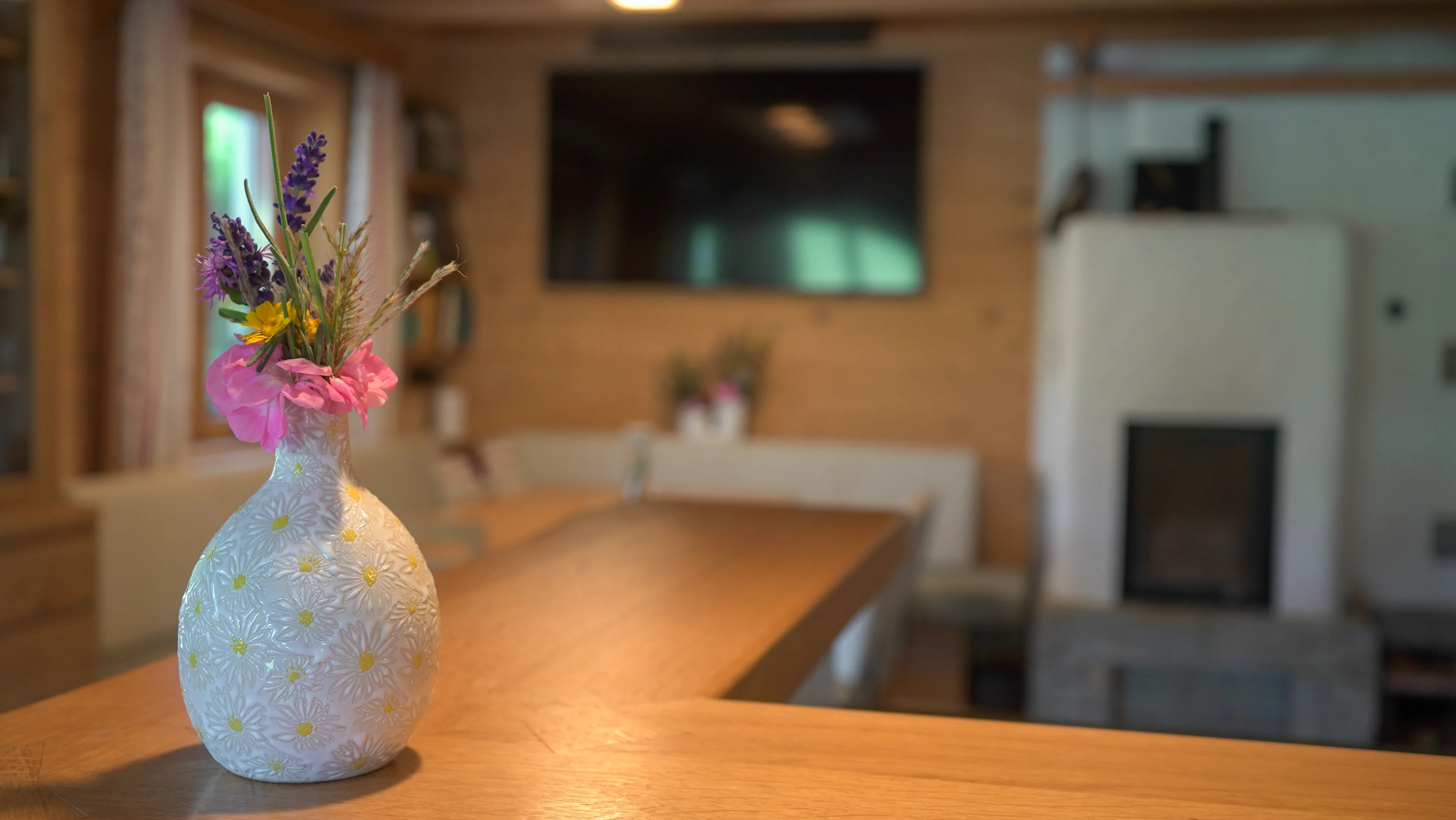 Blumenvase auf einem Holztisch im Seehaus, mit Sitzbank und Kamin im Hintergrund.