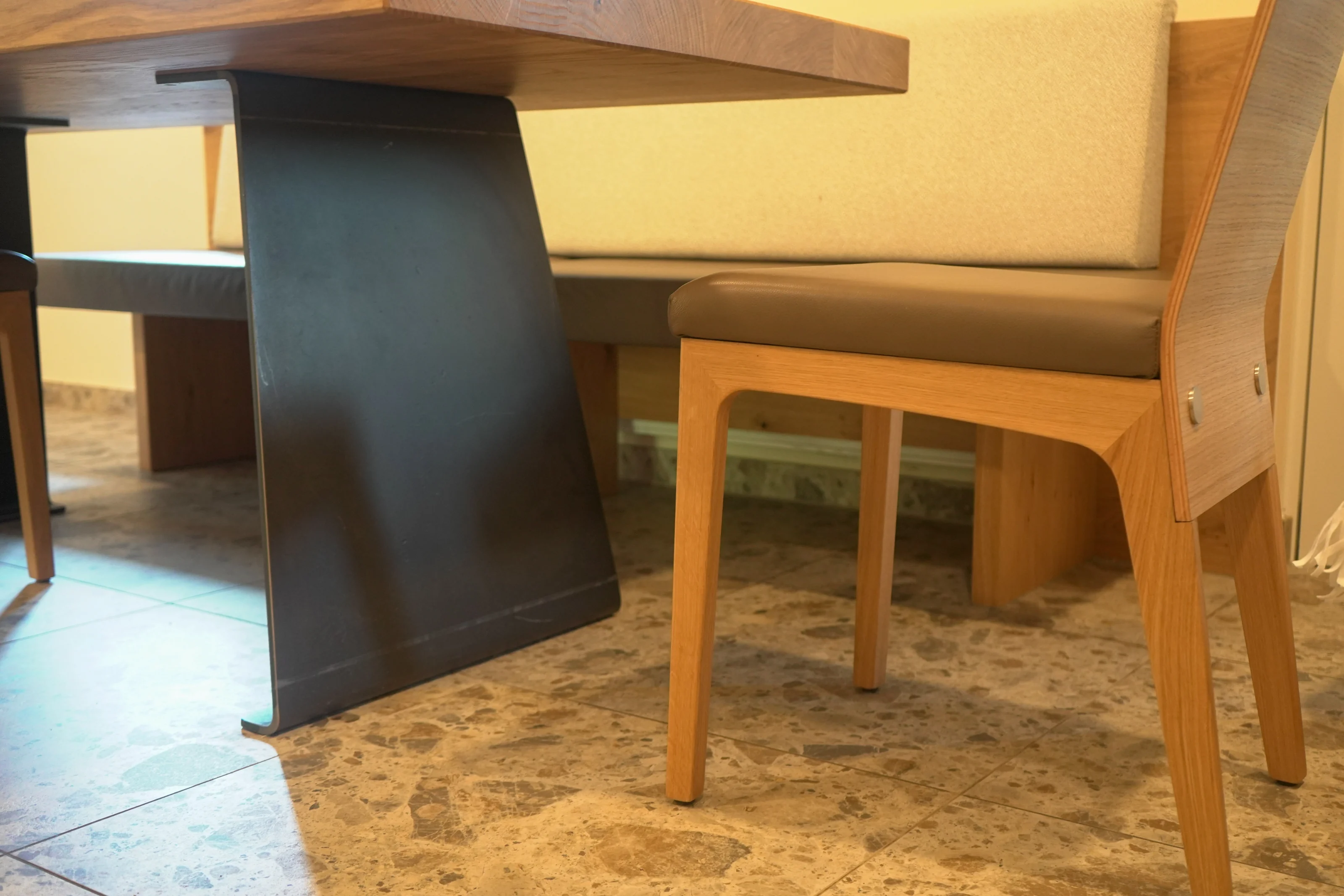 Detail von Holztisch, dunklem Mittelgestell und gepolstertem Stuhl.